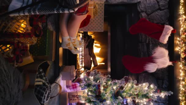 装飾されたXmasの木および暖かい毛布が付いている暖炉の近くの居心地のよいクリスマスのウールの靴下の女性のフィート コピースペースのための垂直ショットクリスマスイブモバイルバックグラウンド 赤いカメラ4Kスローモーションで撮影 — ストック動画
