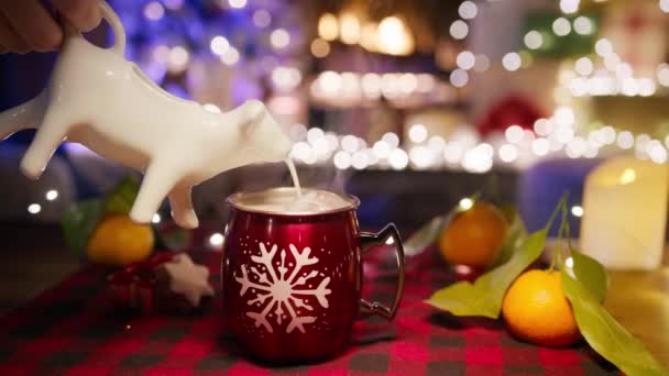 スノーフレークで赤いカップで美味しい甘いホリデードリンク 暖炉の明るいガーランドの平らなナプキンとクリスマスの装飾 ココアに浮かぶホットミルクチョコレート爆弾をこぼす牛の形をしたクリーマーピッチャー — ストック動画