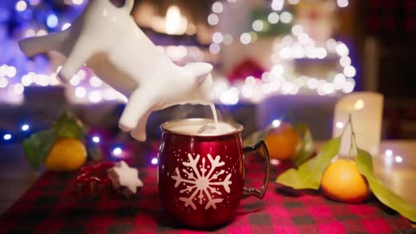 牛の形をしたクリーマーピッチャーから熱いミルクをこぼす女性手は 雪のフレークでココアに浮かぶチョコレート爆弾を溶かします 暖炉の明るいガーランドの平らなナプキンが付いているクリスマスの装飾 — ストック動画