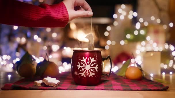 ホリデーで熱いお茶を沸騰する女性はマグをテーマにしました 火の暖炉とクリスマスツリーのぼやけた背景に対して 木のテーブルにスノーフレークが付いた赤い光沢のあるマグでティーパケットを手に入れる — ストック動画