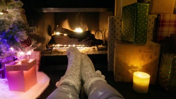 明るい火災で暖まる盗まれた編まれた靴下で足を閉じます 暖炉を燃やし クリスマスイブのリビングルームで輝くクリスマスツリーの隣にある贈り物やプレゼントの大きなヒープの4K映像 — ストック動画