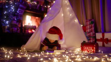 Mutlu Noeller için oyuncak hayal eden tatlı çocuk. Kırmızı Noel Baba şapkalı çocuk, Noel Baba 'ya şömineli bir peri masalı odasında mektup yazıyor. Çadır ışığı altında komik bir çocuk.