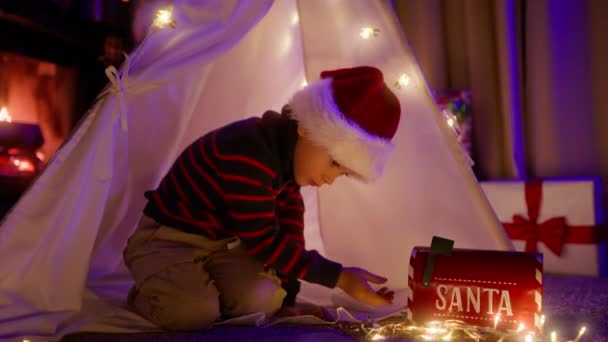 可爱的孩子戴着红色的大圣诞帽 用红色的信箱寄信到北极的圣诞村 快乐的孩子在帐篷里想着珍惜的愿望 在壁炉前享受圣诞时光 男孩梦想着玩具 — 图库视频影像