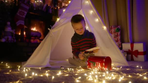 孩子们用红色的信箱寄信给圣诞老人 寄往圣诞村北极 在舒适的壁炉边享受圣诞时光的帐篷里 快乐的孩子在想珍爱的祝福 可爱的男孩梦想玩具4K — 图库视频影像