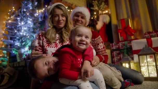 クリスマスの装飾された部屋に座っている小さなかわいい赤ちゃんの娘と幼い息子と幸せな若い両親 冬休みとハッピーニュー2024年を祝うクリスマスイブで楽しい家族 — ストック動画