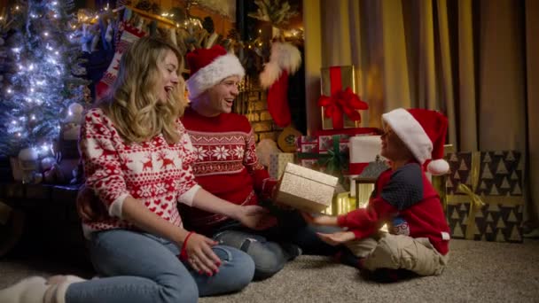赤いサンタの帽子で小さな愛する息子から黄金のギフトボックスを受け取ったハッピーな若い両親は クリスマスのお祝いに楽しい驚きと注意を感謝の意を表します 冬休み おめでとう — ストック動画