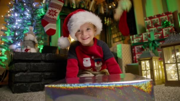光沢のある銀のクリスマスプレゼントボックスに贈り物が何であるかを推測する誘惑で陽気な慎重な小さな男の子の肖像画 かわいい驚いた幸せな顔の子供赤いサンタハットオープニングXmasギフトと興奮した顔 — ストック動画