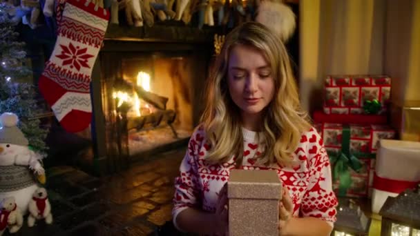 魔法のクリスマスナイトコンセプト4K 居心地の良い装飾された暖炉に座っている白人の美しい女性 幸せな魅力的なブロンドの女性が保持し 黄金の輝く輝く光沢のある赤いカメラで光沢のある贈り物を示しています — ストック動画