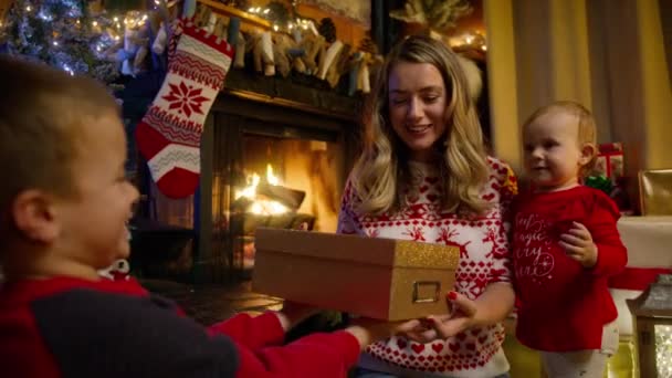かわいい小さな笑顔の少年は クリスマスツリーライトガーランドの近くの暖炉に座っている若い母親と赤ちゃんの妹にスタイリッシュな輝く黄金のクリスマスギフトボックスを与えます 休日にプレゼントを持った幸せな子供 — ストック動画