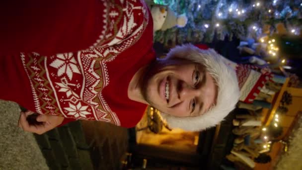 身穿红色圣诞老人帽的快乐年轻人的画像的垂直方向 看着摄像机和录音日志 一个英俊的微笑的家伙在网上拍视频打电话给快乐圣诞致辞 — 图库视频影像