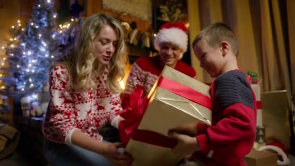 魔法のクリスマスイブに大きな赤い弓で大きな光沢のある黄金の箱でプレゼントを開くことに興奮する幸せな少年 かわいい息子がギフトボックスを揺さぶっている 家族4Kでお祝いの祝日を楽しんでいる興味深い子供 — ストック動画