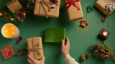 Kadın ve erkek elleri masaya bir sürü renkli hediye kutusu koyuyor. Güzel hediye kutularıyla dolu, yeşil arka planda ve Noel süslemeleriyle dolu bir daire. Genel görünüm hediyeleri arka plan 4K