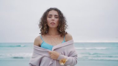 Gri, kasvetli bir günde kumsalda parlak metalik bir bluzla doğal makyajlı güzel kıvırcık kadın portresi. Moda modeli kırmızı kamera. Güzel kız okyanusta poz veriyor, duygusal olarak kameraya bakıyor.