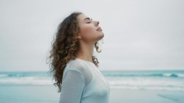 Sakin genç bir kadın derin bir nefes alıyor. Gözleri kapalı meditasyon yapıyor. Arka plandaki okyanus dalgalarına bakıyor. Yan görünüm kızı huzur, zihinsel rahatlama, stres yok.