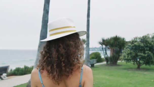 白い帽子の肖像画の若い女性は カーリーヘアが回転し カメラを見て 魅力的に微笑みます 海洋公園の青いドレスの女の子の後ろの赤いカメラ セクシーな汚れた感情的な顔のショット 屋外のライフスタイル — ストック動画