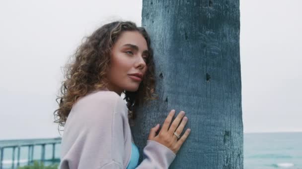 漂亮的卷发女人站在树干上 穿着雅致的羊毛衫 靠近迷人的性感模特 用迷人的蓝眼睛看着相机 自信的女装 时尚美的概念 — 图库视频影像