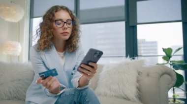 Modern bir evde elinde akıllı telefon ve kredi kartı olan gözlüklü şık bir kadın. Anında kolay ödemeler yapan mutlu bayan alışverişçi, çevrimiçi mağaza uygulamasında müşteri satın alma