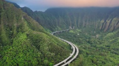 Hawaii 'de gün doğumu sahnesi. Yol tüneli orman dağına doğru uzanıyor. Pembe gün doğumu. Oahu adasındaki dik yüksek dağ sırtı etrafında kıvrılan dolambaçlı otoyol ile sinematik doğa manzarası