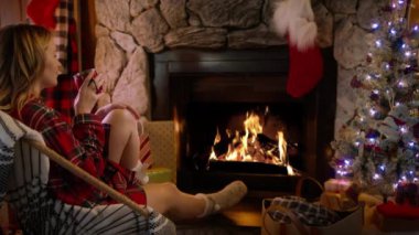 Yanan ateşle rahatlayan ve sıcak çay ya da sıcak çikolatayla ısınan bir kadın. Kış ve Noel tatili konsepti, RED kamera yavaş çekimde. 4K numaralı şöminede yün örgü çoraplı bir kadın.