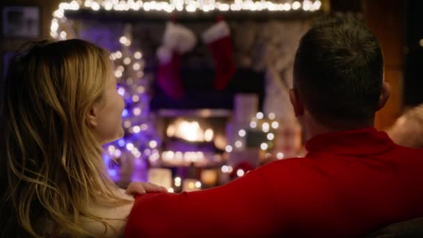 夫妻二人一起坐在客厅的沙发上 观看着燃烧的壁炉 女朋友和男朋友拥抱 拥抱和聊天 圣诞前夕 微笑的女人带着爱意看着她的伴侣 — 图库视频影像