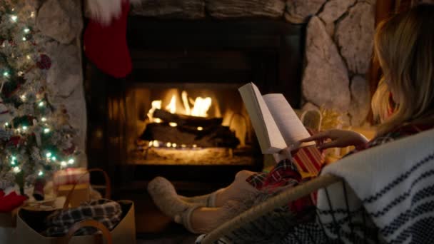 在圣诞节装饰的舒适的壁炉边看书和放松的女人的背景图 松驰的女性坐在椅子上享乐 寒假周末在小木屋里放映机 — 图库视频影像