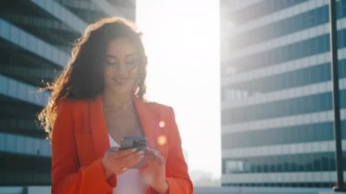 Arka planda altın günbatımı olan akıllı telefondan sohbet mesajı yazan mutlu, genç bir ırk kadını. Gülümseyen iş kadını cep telefonu araması bilgilerini kablosuz internet, online alışveriş 4K üzerinden kullanıyor