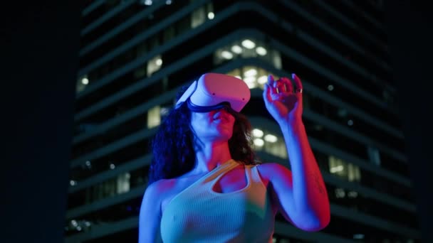 若い女性は 都市の未来的な都市の背景にある青いネオンライトの魔法の雰囲気で仮想または拡張現実のヘルメットを使用しています デジタルインタラクティブアプリの女性Vrヘッドセットユーザー エンターテインメントの未来 — ストック動画