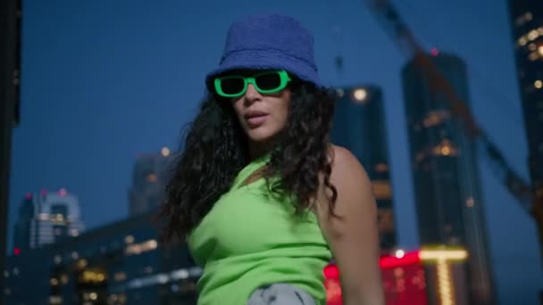 酷酷性感的混血女现代舞选手有节奏地跳4K 身着霓虹灯绿色上衣 头戴紫色斗篷帽的活跃的非洲裔美国黑人女性夜晚在市中心的灯光背景下跳舞 — 图库视频影像