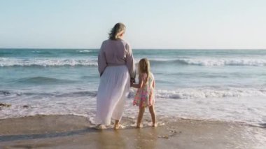 Anne ve kızının el ele tutuşması. Anne ve kız okyanusta durup, altın gün batımında dalgalara bakıyorlar. Ebeveyn ve çocuk, mutlu çocukluk, özgür yaşam tarzı, mutlu aile kavramı