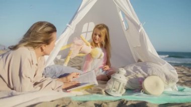 Sarışın genç bir kadın kızı için anılar yaratıyor. Anne, okyanus sahilindeki beyaz çadırda uzanıyor ve küçük kıza kitap okuyor, Ağır çekim 4K. Güzel, tapılası bir anaokulu öğrencisi ailesiyle eğleniyor.