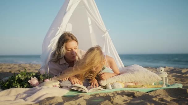 幸せな白人の母親と彼女のかわいい赤ちゃんの女の子は 日没時に海のビーチに設置された白い居心地の良い子供のテントに一緒に横たわって時間を過ごし 本を読んでいます 幸せな家族 遊びの概念による教育 Redカメラ — ストック動画