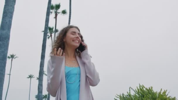 パームツリー付きの公園でスマートフォンで歩くフレンドリーなカーリーレディの低角度ショット 屋外でスマートフォンを話す青いドレスでカジュアルなカーディガンのポジティブな女性 スマイリー 携帯電話 — ストック動画