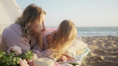 Altın gün batımında okyanus sahilinde güzel, sevgi dolu bir aile, sarışın bir anne ve küçük bir kız birbirlerine bağlanıyorlar. Mutlu ebeveynlik kavramı. Anaokulundaki kız çocuk kitap okuyor tasasız çocuklukta eğleniyor.