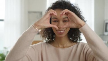 Gündelik bej kazak giyen gülümseyen manken aşkını gösteriyor. Kıvırcık esmer afro saçlı çekici bir kız öğrencinin portresi. Elleriyle kalbi şekillendiriyor ve modern dairede kameraya bakıyor.
