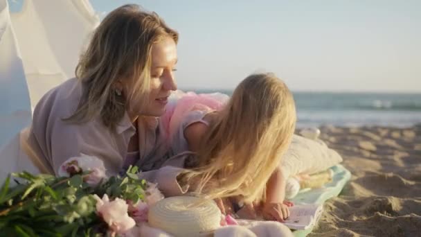 学龄前的小女儿在无忧无虑的童年读书 美丽可爱的家庭 金色的母亲和小女孩在金色的夕阳西下的海滨相拥在一起 快乐父母的概念 — 图库视频影像