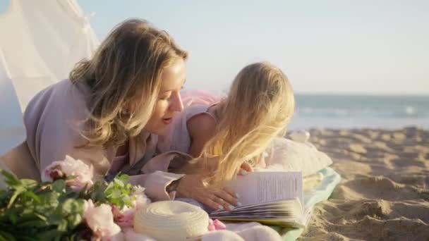 幸せな親のコンセプト 幼稚園児の幼稚園児は 子供の頃から楽しめる本を読んでいました 金の日没の間に海のビーチで結ばれるブロンドの母と小さな少女との美しい愛の家族 — ストック動画