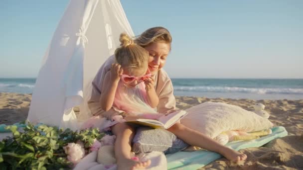 可爱的女孩戴着迷人的粉色心形太阳镜 坐在妈妈的围裙上 夕阳西下时一起看书 母亲和女儿玩得很开心 紧紧地拥抱在一起 美丽的爱家4K — 图库视频影像