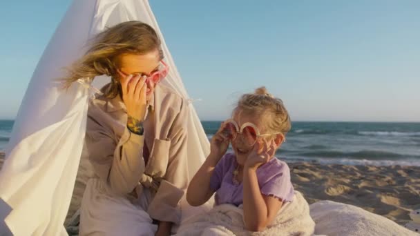 美しい母親は笑う娘を5人で与えています 海のビーチのキャンバステントに座って夏休みの旅行を楽しんでいる少女 一緒に最高の楽しい時間を過ごす母と笑顔の娘 — ストック動画