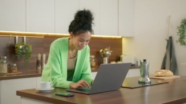 Çift ırklı genç bir kadın dizüstü bilgisayara ve daktiloya bakıyor, çevrimiçi markette bakkal seçiyor, yemek siparişini modern mutfakta yemek masasına yaslanmış vaziyette internet siparişi veriyor.