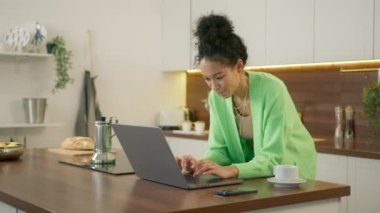 Modern mutfakta, yemek masasına yaslanmış, dizüstü bilgisayara ve daktiloya bakan, çevrimiçi marketten alışveriş yapan, yemek siparişi veren çok ırklı genç bir kadın.
