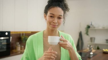 Beyaz bardaktan içen çeşitli modeller, insanların yaşam tarzı 4K. Susamış, gülümseyen 20 'li yaşlarda, çok ırklı bir kadın. Yeni güne bir fincan aromalı kahve içerek başlıyor. Modern mutfağın içinde tek başına duruyor.