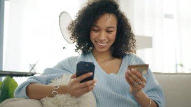 Modern apartmanda cep telefonu ödemelerini kullanarak kredi kartı tutan zarif, çok ırklı genç bir kadın. Gülümseyen koyu tenli güzel kız akıllı telefondan para alıyor.