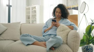 Gülümseyen Afro-Amerikan tutkulu kadın elinde akıllı telefon tutuyor. Çift ırklı kız cep telefonu kullanıyor. Evde gün ışığında oturup sosyal medya izliyor. Gerçek insanların yaşam tarzı akıllı telefon eğlencesi 4K