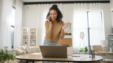 Genç Afro-Amerikan bayan girişimci dizüstü bilgisayarda çalışıyor ve dahili ofisteki akıllı telefondan müşteriyle veya ortağıyla konuşuyor. Pozitif küçük işletme sahibi yönetim projesi ve çoklu görev 4K