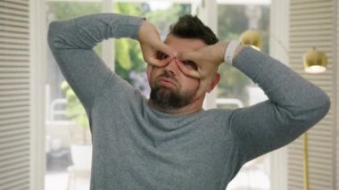 Modern apartman arka planında parmakları olan süper kahraman taklidi yapan şık saçlı yetişkin bir adamın portresi. Komik adam etrafta dolanıyor, komik çocuksu suratlar yapıyor 4K