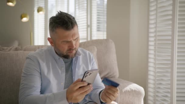 ハッピーハンサムな男は 現代のリビングルームでソファーに座って笑顔でスマートフォンとクレジットカードでオンラインショッピング 金融とモダンなテクノロジーコンセプト 銀行モバイルサービス モダンなライフスタイル — ストック動画