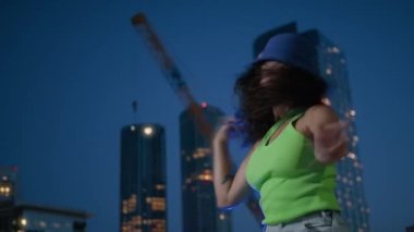 Gülümseyen seksi kız ellerini hareket ettiriyor ve uzun kıvırcık saçlarını 4K yapıyor. Kozmopolit modern şehrin gece caddesinde dans eden güzel ve mutlu Afro-Amerikan kızının 4K şehir arka planında portresi.