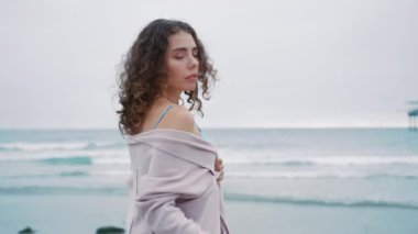 Moda modeli kırmızı kamera. Güzel kız okyanusta poz veriyor, duygusal olarak kameraya bakıyor. Güzel kıvırcık kadın portresi, doğal makyajlı, parlak metalik düz bluzlu kasvetli gri bir günde kumsalda.