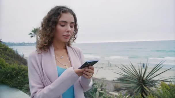 Okyanus Arka Planında Akıllı Telefonuyla Yürüyen Sevimli Kıvırcık Kız Dışarıda — Stok video