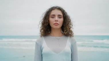Kıvırcık saçlı, harika bir bayan 4K kamerasına bakıyor. Pastel gri, taze gökyüzü arkaplanlı kameraya bakan kızın portresi. Genç bir kadınla doğal güzellik ya da moda reklamı.